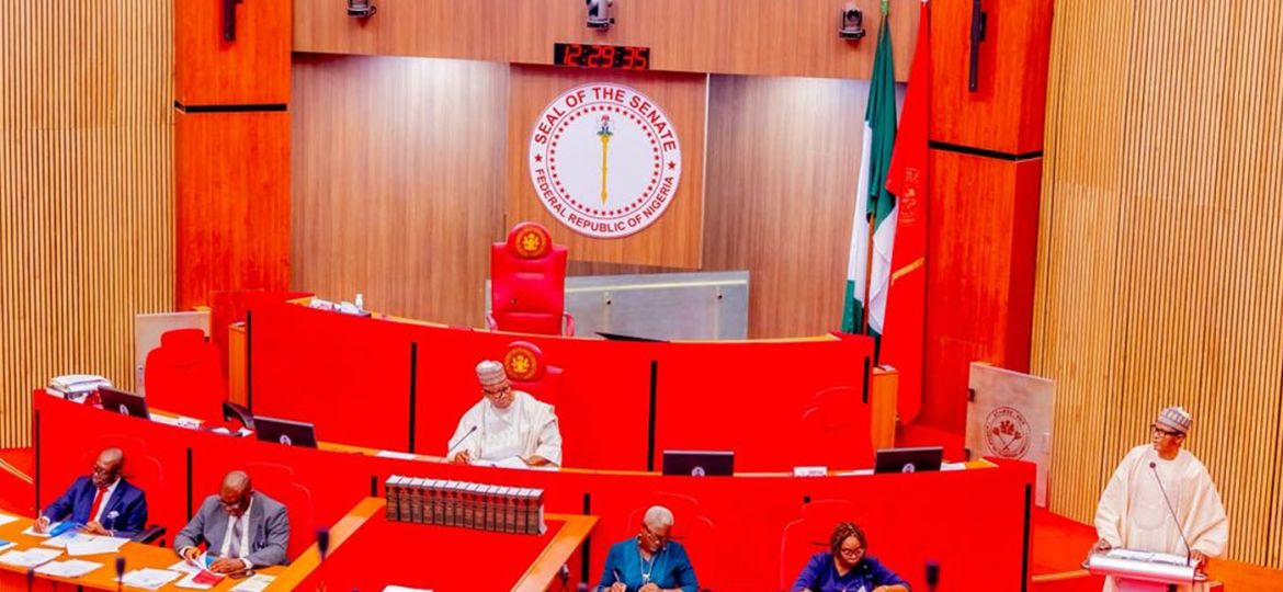 Senate chamber with red desks and seats, Nigerian seal behind the podium, and officials seated and writing at their desks.