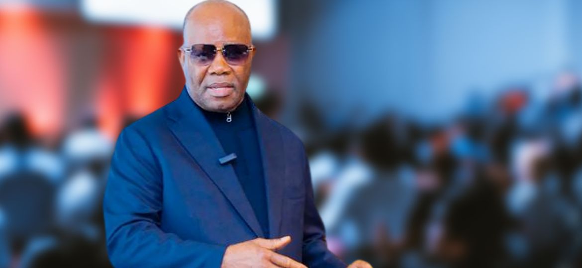 Male presenter in a blue suit and sunglasses delivering a talk to a blurred audience at an event.