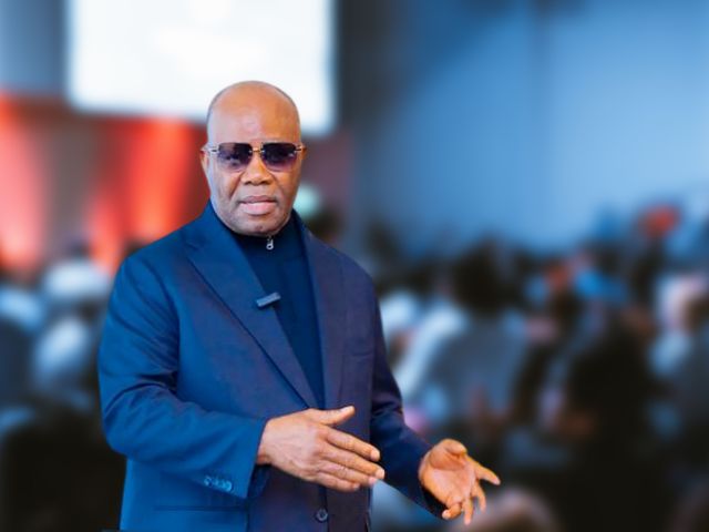 Male presenter in a blue suit and sunglasses delivering a talk to a blurred audience at an event.