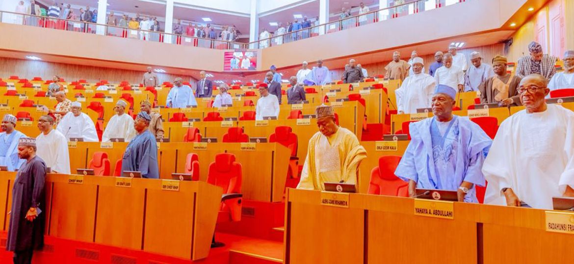 Large conference hall with orange desks and red seats; many men in traditional attire standing between desks in a parliamentary chamber.