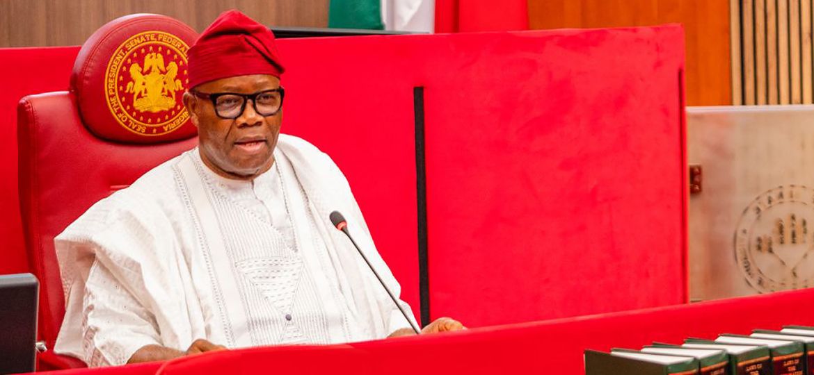 Nigerian official in white agbada and red cap sits at a red bench with a microphone in front of him, behind seals and flags in a formal chamber.