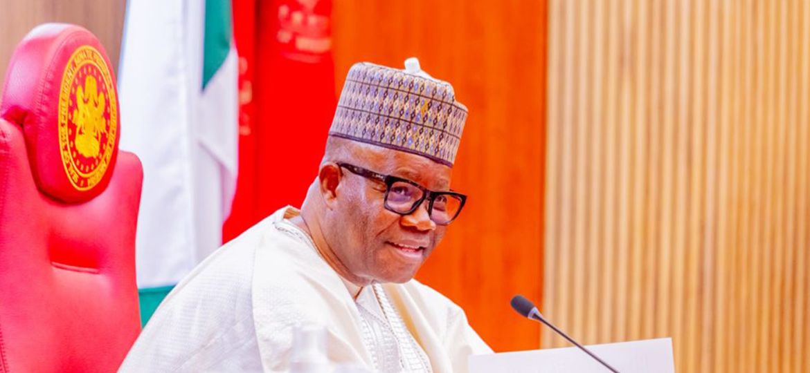 Nigerian official wearing a patterned cap and glasses, seated at a desk with a microphone and papers in front.
