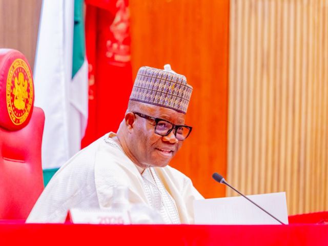 Nigerian official wearing a patterned cap and glasses, seated at a desk with a microphone and papers in front.