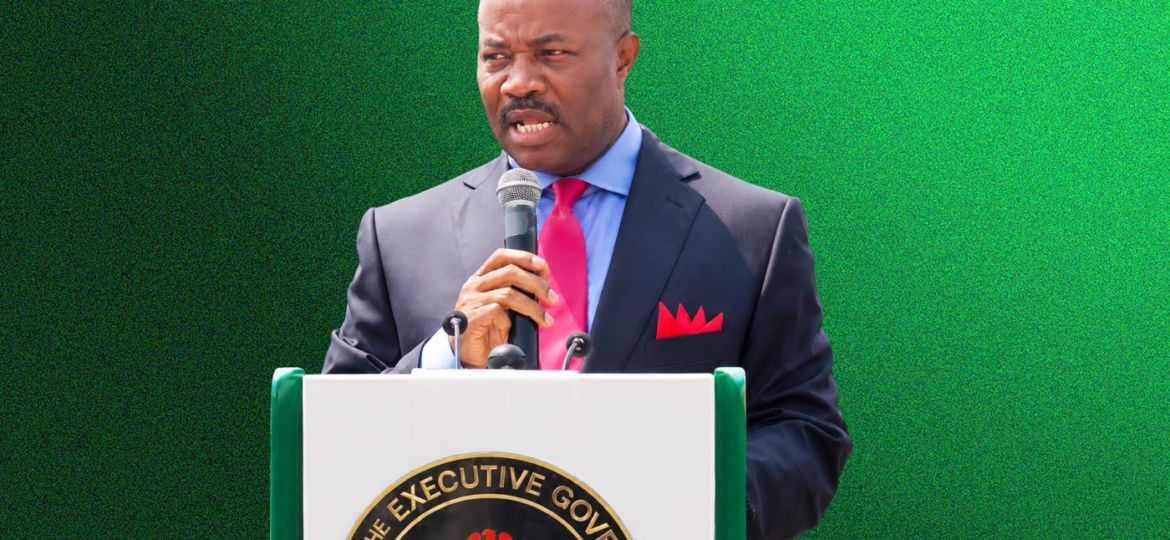 A man in a dark suit and pink tie speaking into a microphone at a podium with a state seal, against a green background.